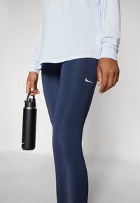 Námornícke legíny s bielym logom Nike, v kombinácii s svetlým modrým tričkom s dlhým rukávom. V pravej ruke je čierna kovová fľaša na vodu.