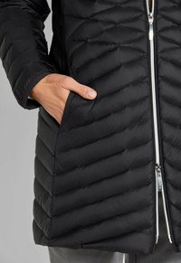 Hand in de zak van een zwarte gewatteerde jas met diagonale stiksels en een zilveren voorrits, gedragen over grijze broeken.