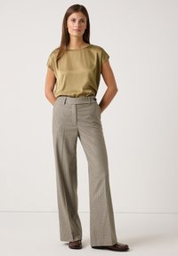 Olivenfarbene Satinbluse mit kurzen Ärmeln, kombiniert mit hochtaillierten, plissierten Karohosen in Beige- und Brauntönen. Braune Schuhe.