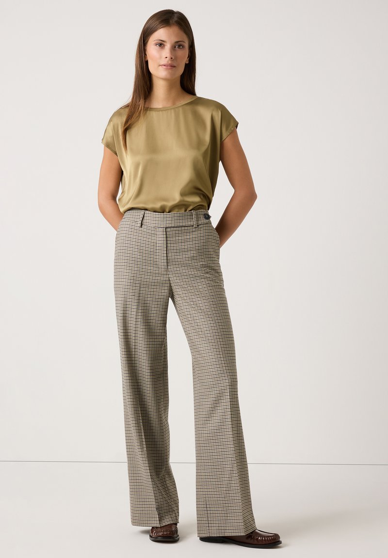 Olivenfarbene Satinbluse mit kurzen Ärmeln, kombiniert mit hochtaillierten, plissierten Karohosen in Beige- und Brauntönen. Braune Schuhe.