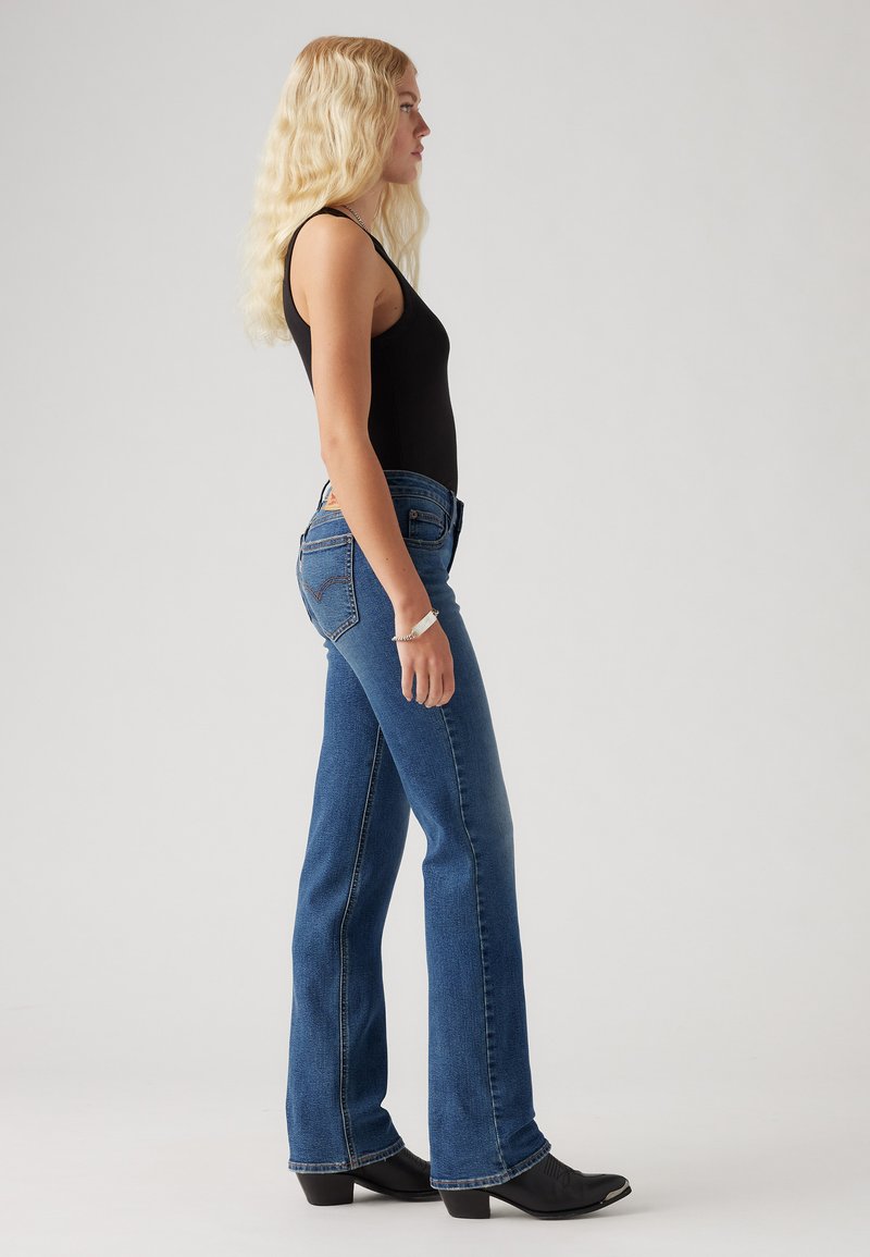 Jean flare en denim bleu taille haute avec un design classique à cinq poches et des coutures contrastées, associé à un haut sans manches noir et des bottes noires.