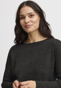 Pull en maille texturée vert foncé avec col rond et manches longues, présentant un léger détail de couture sur le devant.