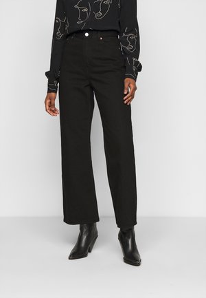 Persoon draagt zwarte high-waist broek met rechte pijpen, zwarte enkellaarzen met puntige neuzen en een zwart overhemd met witte abstracte gezichtslijnkunst.