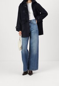 Mujer vistiendo un abrigo negro con capucha, una blusa blanca, jeans azules de pierna ancha, botas marrones hasta el tobillo y sosteniendo un pequeño bolso blanco.