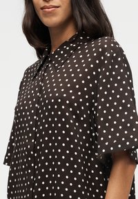 Femme portant une chemise noire à manches courtes boutonnée avec des pois blancs, montrant le col et une partie de son visage et de son bras.