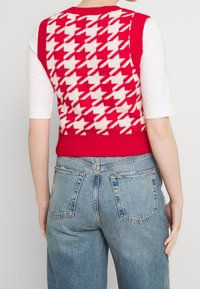 Czerwona i kremowa kamizelka w wzór pepitki noszona na białej bluzce, zestawiona z jasnoniebieskimi, wysokim stanem dżinsami. Dzianinowa faktura z prążkowanym wykończeniem na dole.