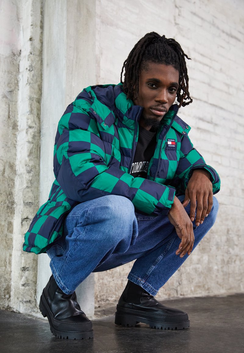 Zielono-granatowa pikowana kurtka w kratę, wyposażona w zamek błyskawiczny i dwie boczne kieszenie. Ciemnoniebieskie dżinsy i czarne, masywne botki dopełniają całość.