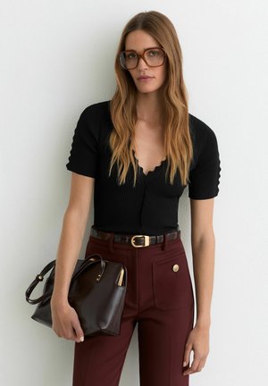 Pika helepruuni juustega naine, kellel on suured pruunid prillid, must lühikeste varrukatega särk, burgundia püksid ja käes tumepruun käekott, taustaks valge sein.