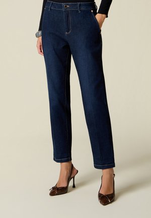 Person wearing dark blue straight-leg jeans with front pockets and brown high-heeled slingback shoes, standing on a plain background.