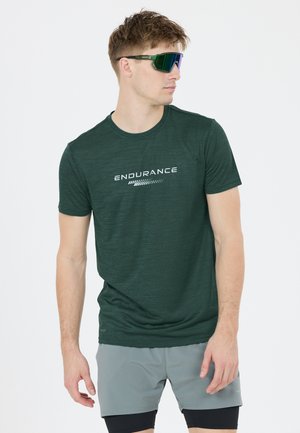 Jonge man met een donkergroen "Endurance" T-shirt, grijze korte broek, zwarte legging en grote reflecterende sportzonnebril, kijkt opzij.