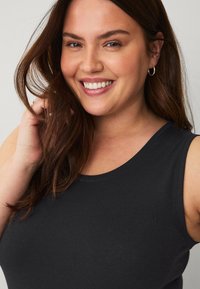 Zwarte mouwloze tanktop met een ronde halslijn, een soepele stof en minimale stikseldetails, gedragen door een persoon met lang bruin haar.