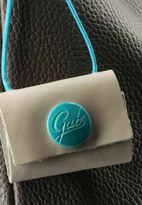 Cartera de cuero beige con un cierre circular turquesa brillante que presenta el texto "Gabs". Incluye una correa trenzada azul. Fondo texturizado.