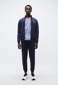 Ναυτικό μπλε φόρμα με ζακέτα με φερμουάρ, ταιριαστό παντελόνι και ανοιχτό γαλάζιο μπλουζάκι. Διακοσμημένο με λογότυπο. Φορώντας το με μαύρα και άσπρα αθλητικά παπούτσια.