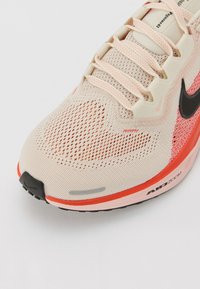 Bež mrežasta atletska cipela s koraljnim akcentima, crnim Nike logom i teksturiranim gornjim dijelom. Ima podloženu jedinicu s oznakom "AIR ZOOM".