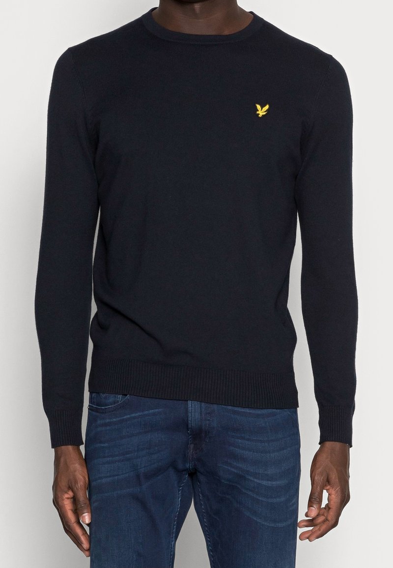 Maglione a maglia blu navy con scollo e orlo a coste. Presenta un piccolo logo giallo sul petto sinistro. Abbinato a jeans blu.