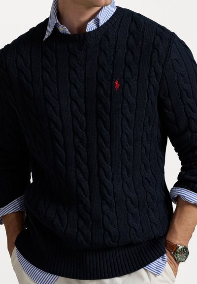 Mörkblå kabelstickad tröja med rund halsringning och en röd broderad logotyp. Blårandiga skjortärmar syns vid ärmarna.