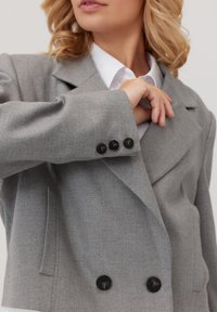 Szary dwurzędowy żakiet z szerokimi klapami, teksturowaną tkaniną i czarnymi guzikami. Posiada boczne kieszenie oraz ogólnie dopasowany krój.