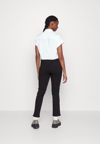 Camisa branca de mangas curtas com punhos dobrados, combinada com calças ajustadas pretas e sapatos cinzentos, vista pela parte de trás contra um fundo simples.