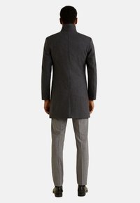 Homme vu de dos portant un long manteau gris foncé, un pantalon gris clair et des chaussures habillées noires, debout devant un fond blanc uni.