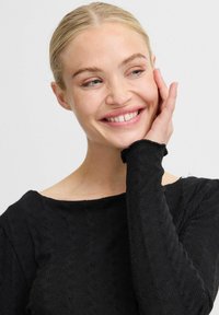 Pulover negru tricotat cu un model texturat, guler larg și mâneci lungi cu margini desfăcute. Modelul zâmbește și se atinge ușor de față.