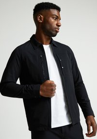 Melns žakets ar rāvējslēdzēju, divām priekšējām kabatām un apkakles izgriezumu, uzvilkts virs baltas T-krekliņa ar garām piedurknēm.