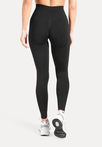 Schwarze Leggings mit hohem Bund, strukturiertem Muster und glatter Oberfläche. Kombiniert mit weißen Sportschuhen, die Details an Knöchel und Ferse betonen.