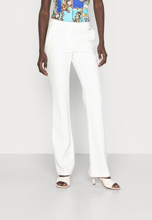 Vrouw die witte, hoog getailleerde aansluitende broek draagt, een blauw patroonloos mouwloos top en witte open teen sandalen met hakken, staand tegen een effen achtergrond.