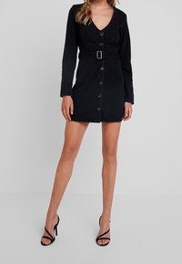 Svart denimklänning med långa ärmar, V-ringning, framknappar och midja med bälte. Har en figursydd siluett och en utsvängd kjol.