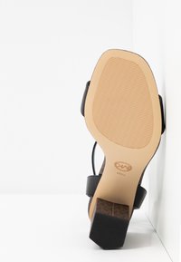 Sandale avec des brides noires et une semelle beige texturée, reposant sur son talon contre une surface blanche en angle.