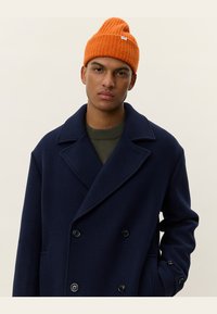 Abrigo de lana azul marino de doble botonadura con botones negros, superpuesto a un suéter de punto verde olivo y combinado con un gorro de canalé naranja.