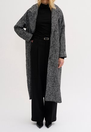 Femme portant un long manteau texturé noir et blanc sur un col roulé noir, un pantalon à jambes larges et des chaussures noires pointues, debout avec une main dans la poche.