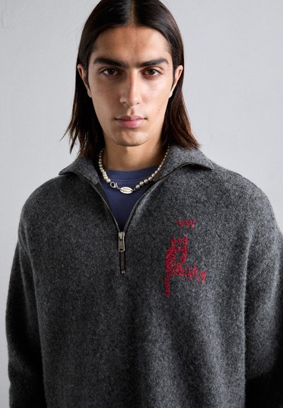 Grauer Wollzipper-Pullover mit einem roten gestickten Katzendesign, Kragen und Zip-Verschluss. Getragen über einem blauen Hemd und einer Perlenkette.