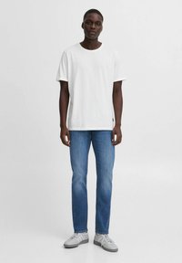 Camiseta blanca de algodón con cuello redondo, combinada con jeans azules. El modelo lleva zapatillas grises con detalles blancos. Diseño simple y casual.