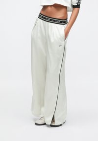 Γυναικεία λευκά παντελόνια Nike σε άνετη γραμμή, με μαύρη μέση και πλευρικές λεπτομέρειες, διαθέτουν τσέπες και απαλή, ελαφριά ύφανση.
