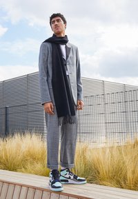 Šedý kostkovaný oblek s černým šátkem, bílou košilí a vícero barevnými teniskami. Model stojí na dřevěné platform�ě s pozadím trávy.