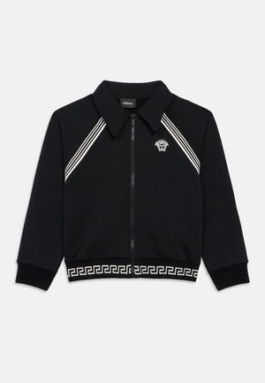 Sort jakke med glidelås med hvit Medusa-logo på brystet, hvite diagonale striper nær skuldrene og gresk nøkkelmønster langs kanten.