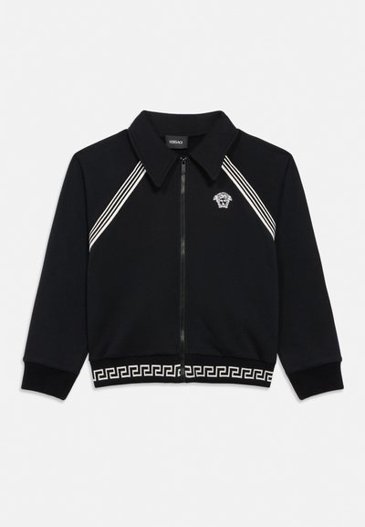 Mustast tõmblukuga jakk valge Medusa logoga rinnal, valged diagonaalsed triibud õlgade lähedal ja kreeka võtmemuster ääreäärsel osal.