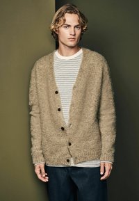 Beige strikket cardigan med en blød, fuzzy tekstur. Den har en knaplukning foran og ribbede manchetter, båret over en stribet hvid og sort skjorte.