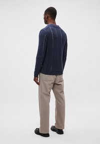 Tengerészkék kötött pulóver, gallérral, függőleges nyitott mintákkal, bézs nadrággal és fekete cipővel párosítva, hátulról nézve.