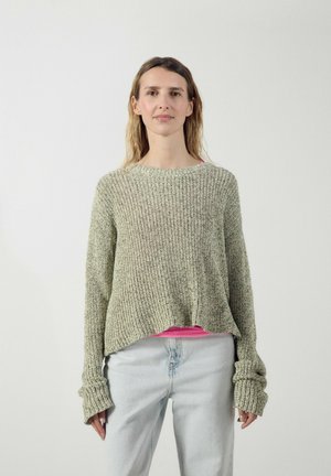 Kvinde står vendt fremad iført en løs grå strikket sweater over en lys pink skjorte og lyseblå jeans mod en ensfarvet lys baggrund.