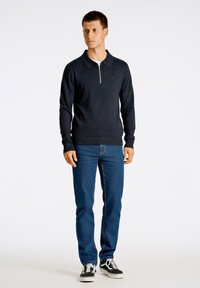 Mörkblå långärmad polo med dragkedja i kragen, ribbade ärmslut och nederkant. Kombinerad med blå denimjeans och svarta sneakers med vita sulor.