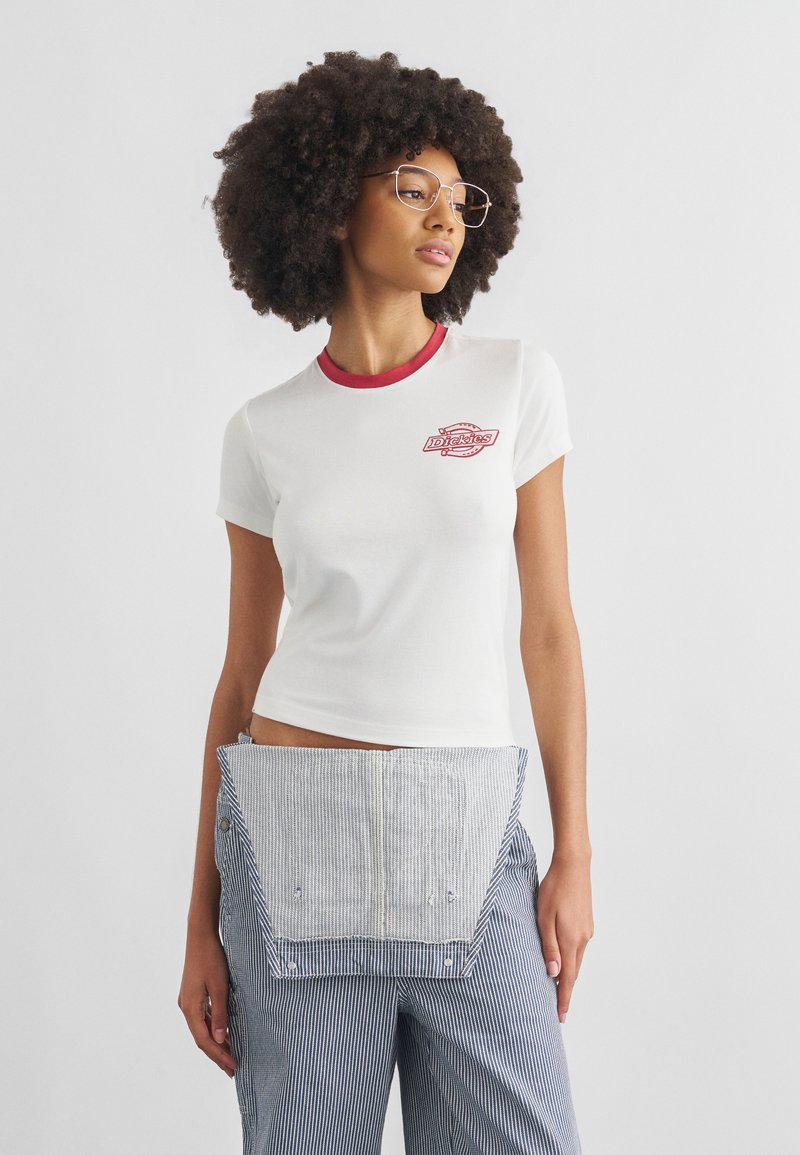 Νεαρή γυναίκα με αφροαμερικάνικη κόμμωση που φοράει λευκό μπλουζάκι Dickies με κόκκινο γιακά και ριγέ παντελόνια με προσαρτημένο πάνελ ποδιάς.