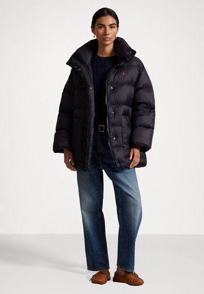 Zwarte pufferjack met een hoge kraag, ritsluiting en drukknoopjes. Gedragen over een donkere gebreide trui en gecombineerd met blauwe jeans.