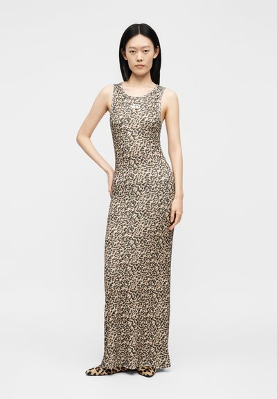 Γυναίκα με ίσια μαύρα μαλλιά που φοράει μακριά αμάνικη φόρεμα με σχέδιο λεοπάρ και ταιριαστά παπούτσια με σχέδιο λεοπάρ, στέκεται με το ένα χέρι στη μέση.