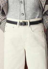 Jeans beige a vita alta con cuciture chiare e cinque tasche abbinati a una cintura in pelle nera con fibbia argentata. Maglione grigio a trama.