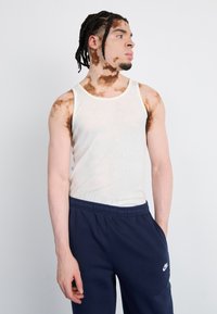 Valkoinen ribattu hihaton toppi yhdistettynä tummansinisiin verkkareihin, joissa on pieni valkoinen Nike-logo vasemmassa lantiovyöhykkeessä. Silkkinen kangas, rento malli.