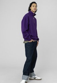 Sweat-shirt violet à col montant, fermeture éclair à l'avant, associé à un jean denim ample. Baskets blanches avec des rayures noires et ourlets de jean retroussés.