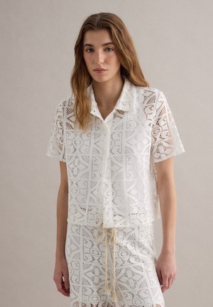 Femme portant une chemise blanche en dentelle à manches courtes boutonnée et une jupe taille haute assortie avec cordon, debout devant un fond uni.