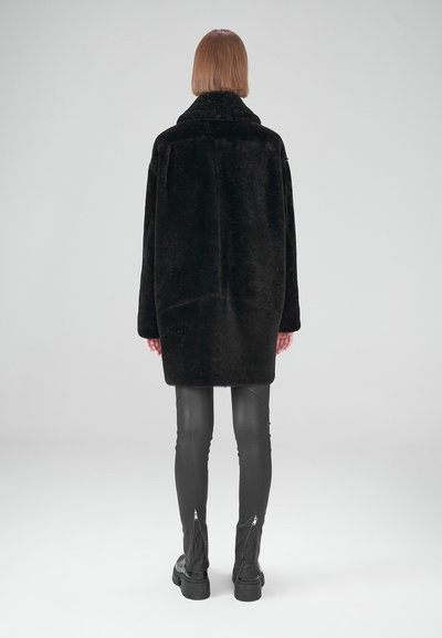 Schwarzer Kunstfellmantel mit hohem Kragen, lockerer Passform und langen Ärmeln, kombiniert mit eng anliegenden schwarzen Lederhosen mit Reißverschlüssen an den Knöcheln.