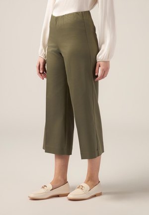 Pantaloni cropped verde oliva con una texture liscia, vita elastica e una sottile apertura laterale, abbinati a mocassini beige con un accentuazione dorata.